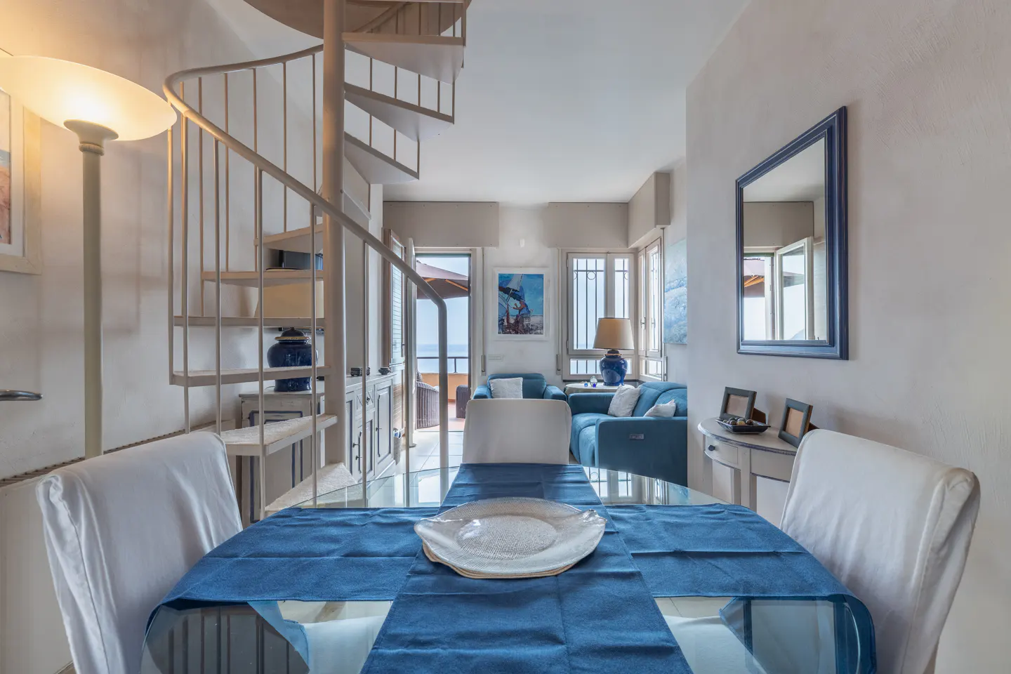 Interior view of a dining area with a glass table, blue runner, white chairs, and a spiral staircase leading to a living room with ocean views.