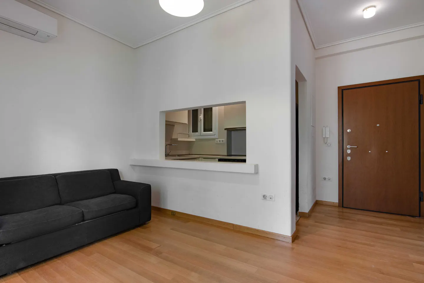 A bright living room with a dark gray sofa, light wood floors, and a view into the kitchen through a rectangular opening in the white wall. A brown door is on the right.
