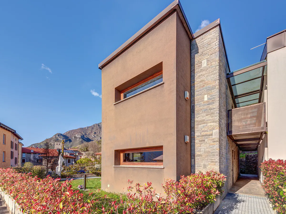 Modern home exterior with brown stucco, stone accents, and a glass-covered balcony. Red bushes line the front, with a mountain backdrop under a blue sky.
