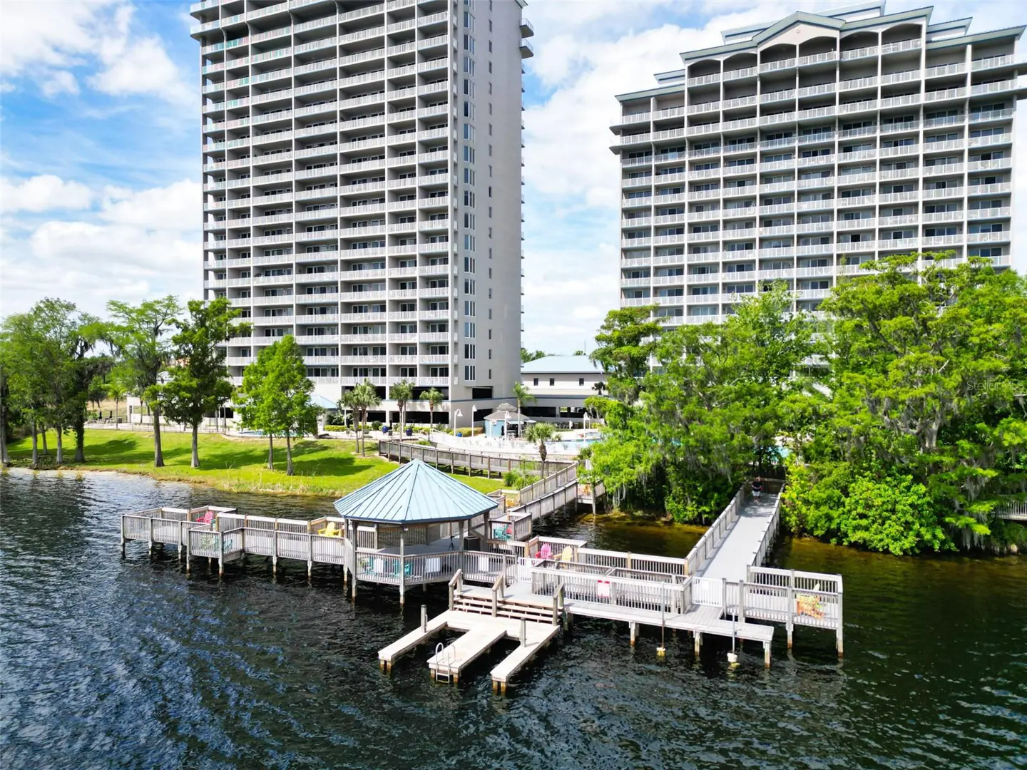 Aerial view of two tall condo buildings behind a lake with a wooden dock and gazebo.