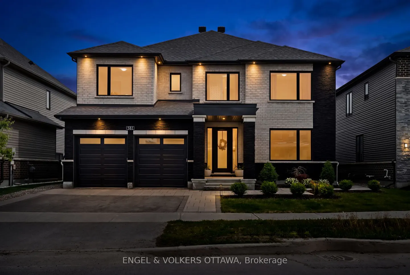 Two-story modern home with black and white brick exterior, double garage, and warm interior lighting at twilight.