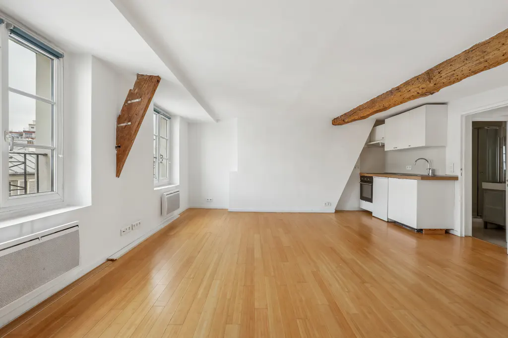 Bright, empty apartment with wood floors, white walls, exposed beams, and a small kitchen area. Windows overlook a city.