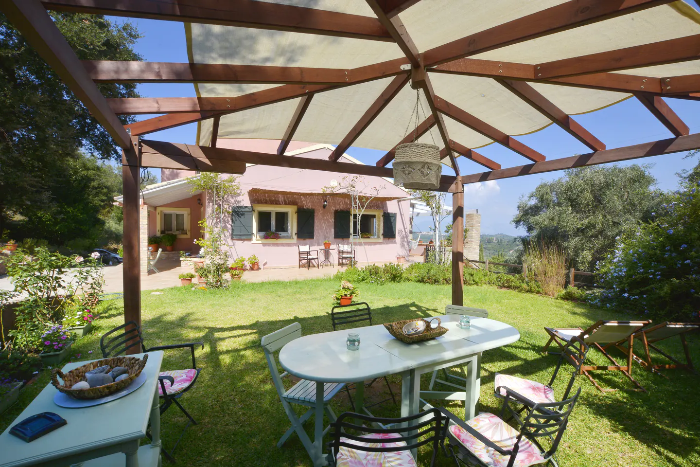 Outdoor dining area with white table and chairs under a pergola, a pink house in the background, and green lawn.