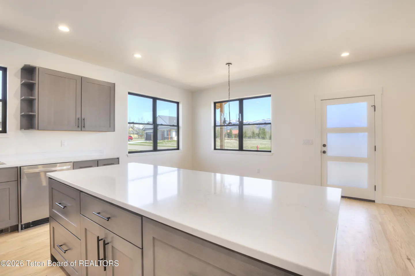 Bright kitchen with gray cabinets, white countertops, and stainless steel appliances. Windows offer views of the outdoors.