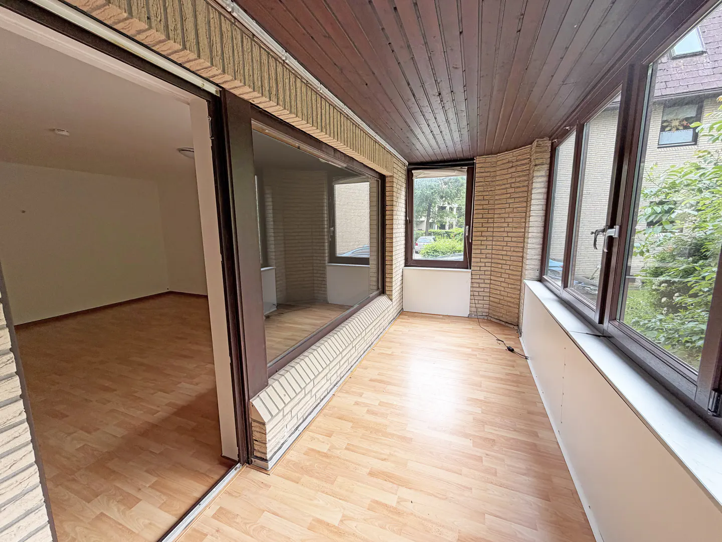 Sunroom with light wood flooring, brick walls, dark wood ceiling, and large windows overlooking a garden.