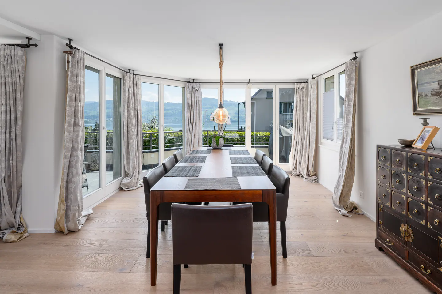 Bright dining room with a long wooden table, gray chairs, and a view of a lake through large windows with patterned curtains. A dark cabinet sits on the right.