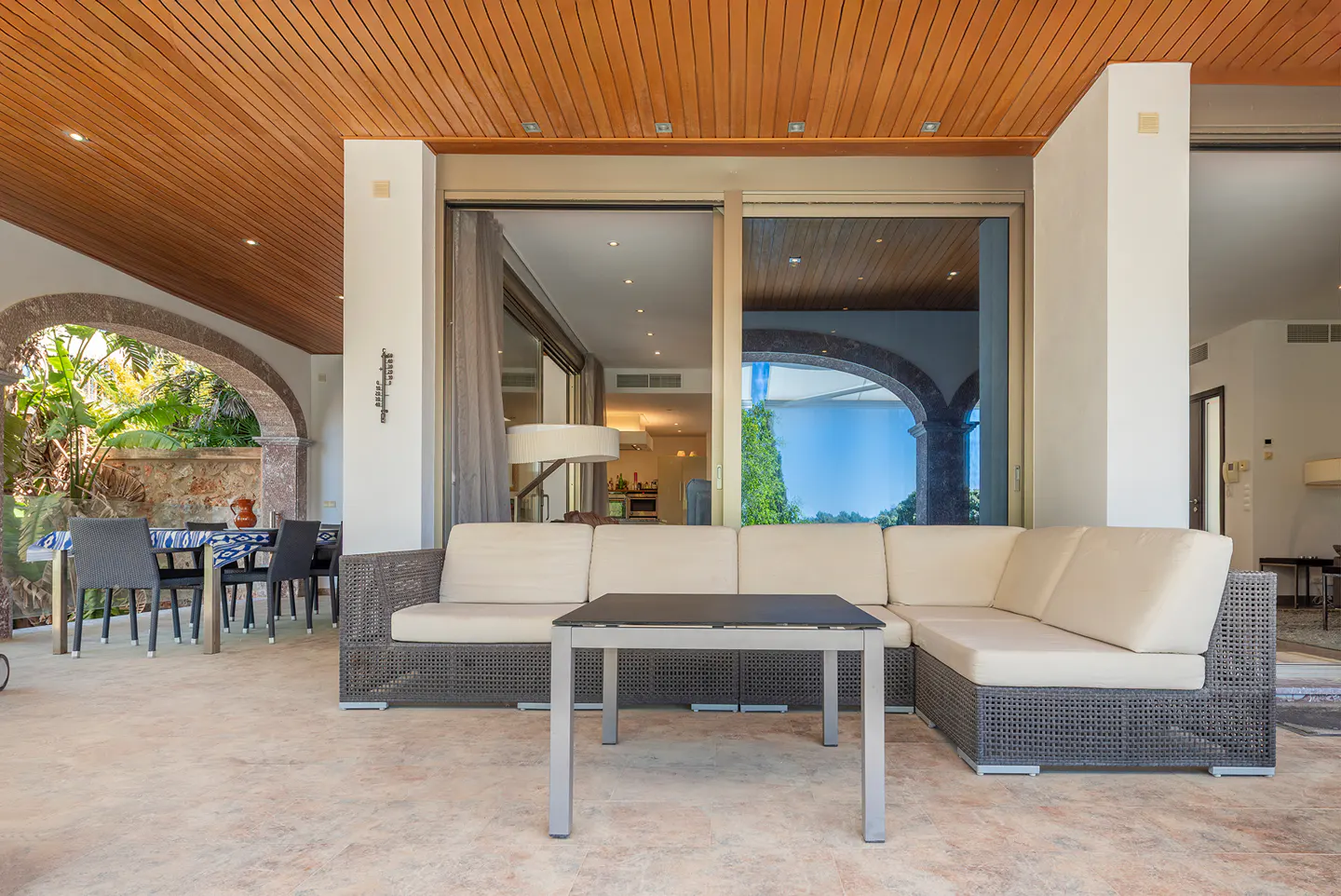 Outdoor patio with wicker sofa, table, and dining set under a wood ceiling. Arched doorway leads to lush greenery. Sliding glass doors open to the interior.