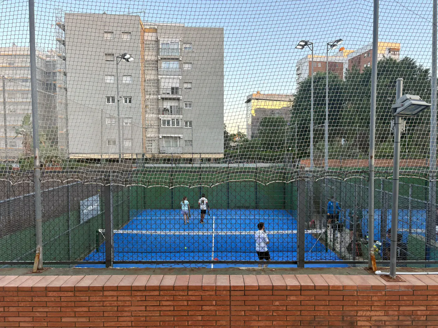 Outdoor blue paddle tennis court with players, viewed through a net fence, with buildings in the background.