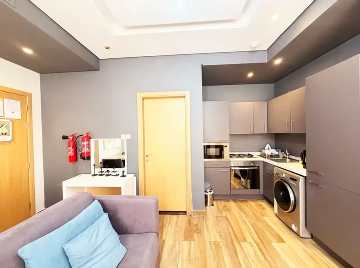 Interior view of a modern apartment with gray walls, wood floors, and a lavender sofa with blue pillows. A gray kitchen with appliances is visible.