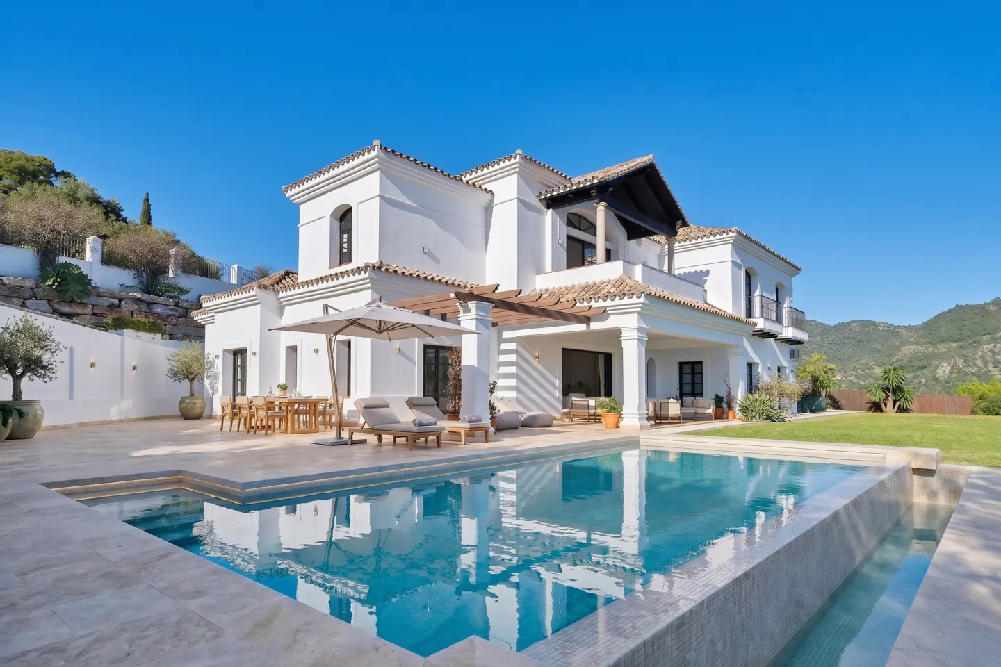 Luxury white villa with a blue tiled pool, lounge chairs, and outdoor dining under a beige umbrella on a sunny day.