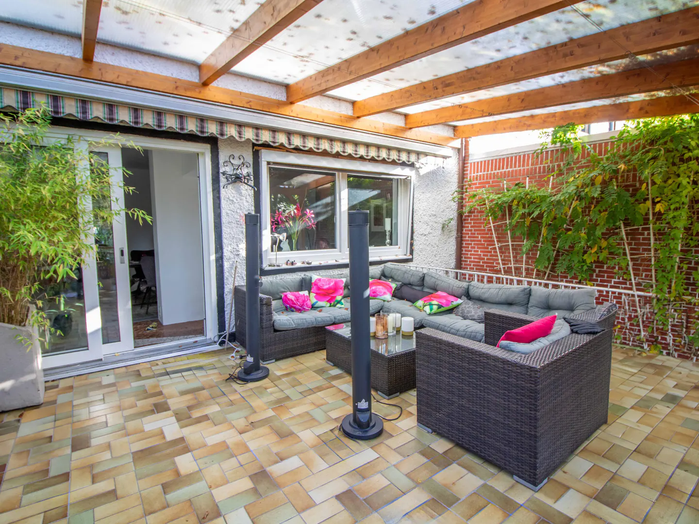Outdoor patio with wicker furniture, gray cushions, and pink pillows. A glass table holds candles. A brick wall with vines is in the background.