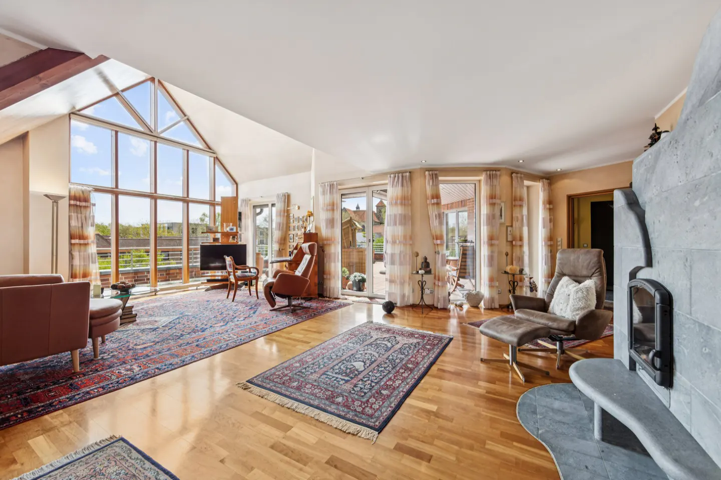 Bright living room with hardwood floors, large windows, and a stone fireplace. Furniture includes leather chairs, a sofa, and patterned rugs.