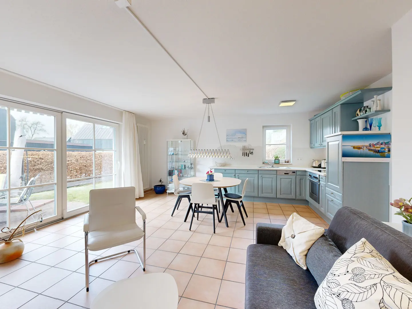 Bright, open-plan living space with a gray sofa, white chairs, and a round dining table. The kitchen features light blue cabinets. Large windows offer a view of the garden.