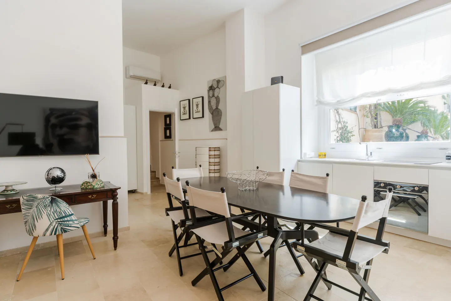 Bright, modern dining area with a dark wood table, canvas chairs, and a TV on the wall. A window overlooks a lush garden.