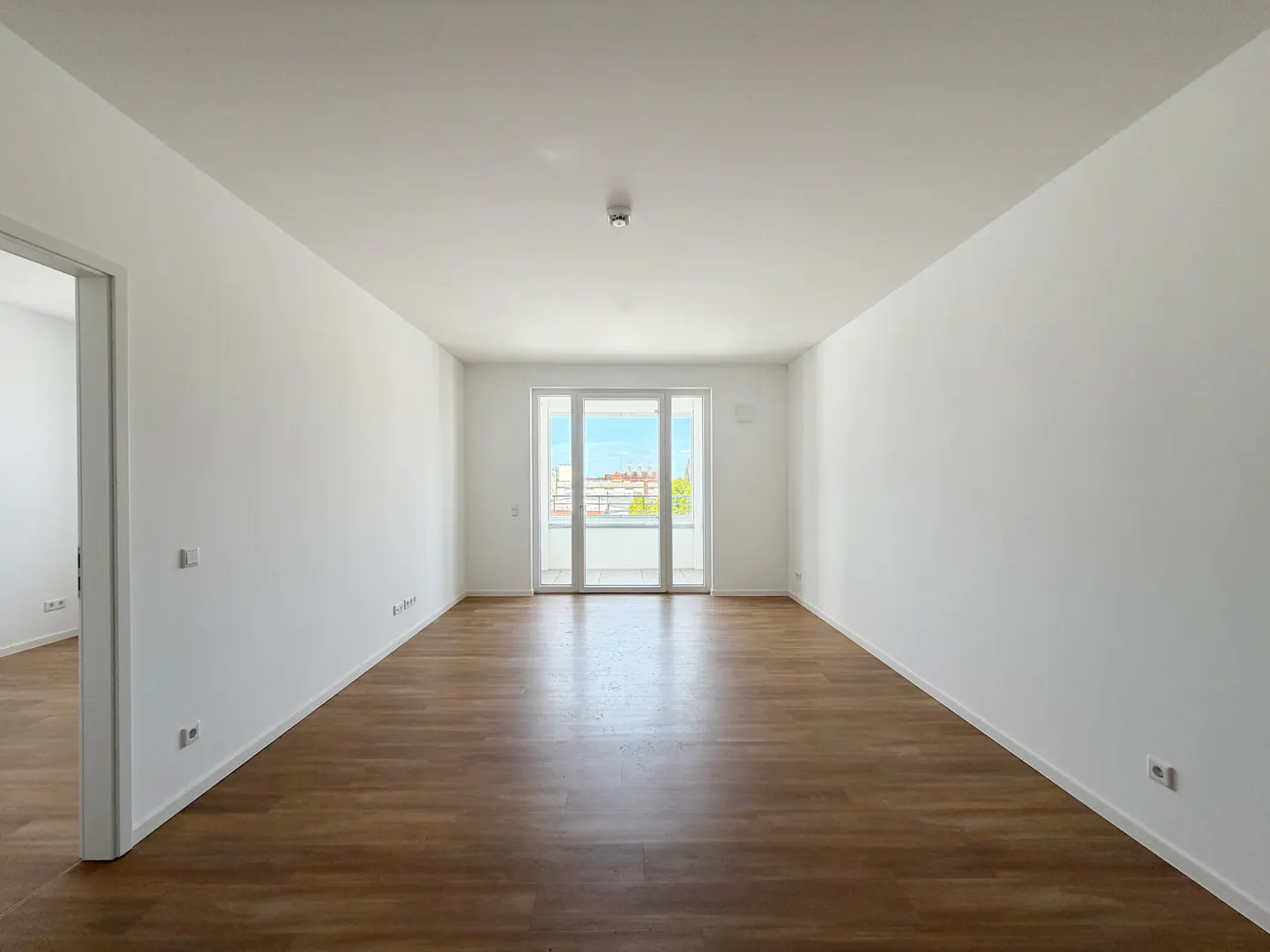 Bright, empty room with wood floors and white walls. A glass door leads to a balcony with a city view.