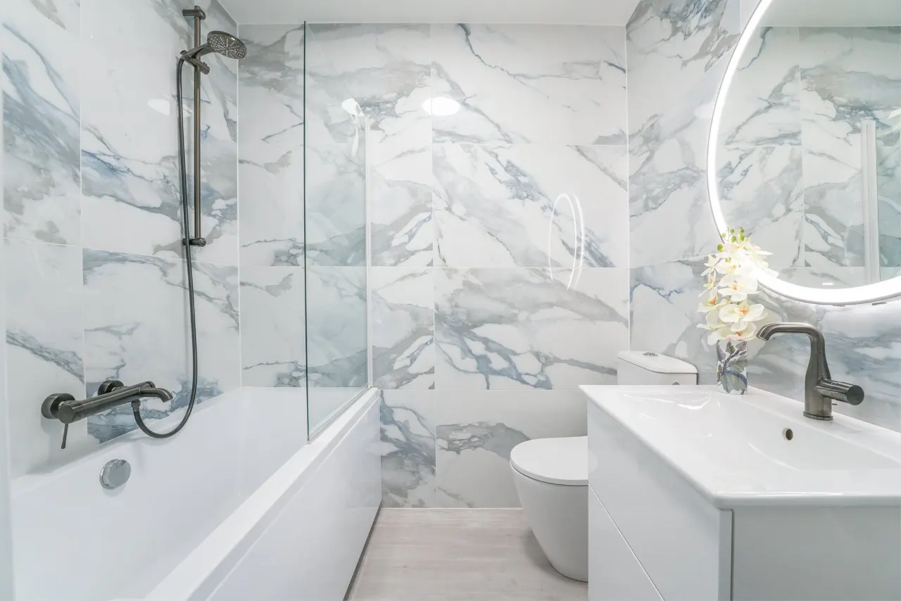 Modern bathroom with white marble-patterned walls, a glass-enclosed tub, a white toilet, and a vanity with a backlit mirror.