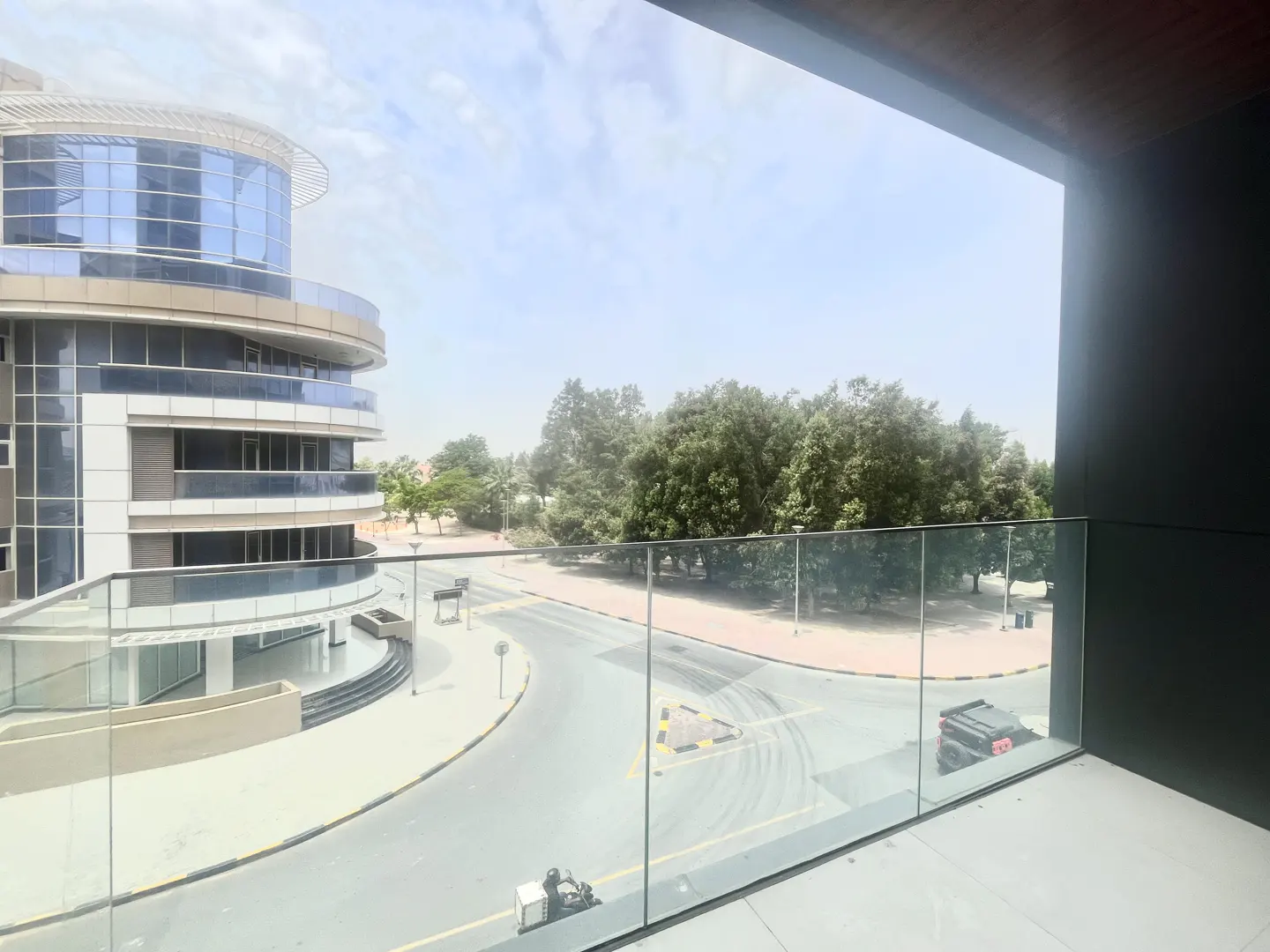 View from a balcony with glass railings overlooking a street, trees, and a modern building on a sunny day.