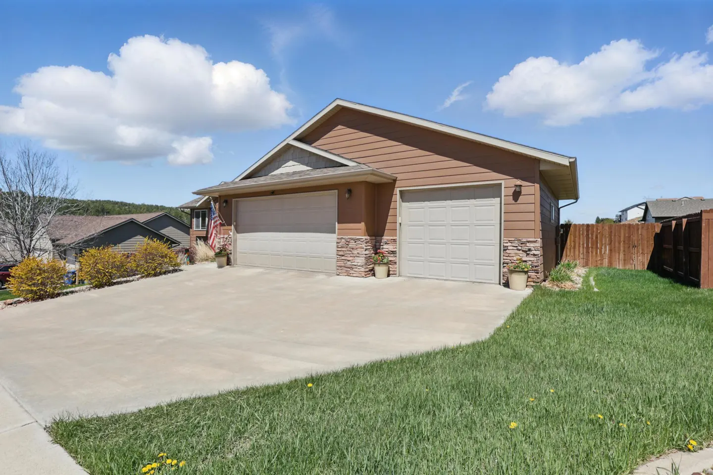 Brown house with a three-car garage, stone accents, concrete driveway, and green lawn under a blue sky with clouds.
