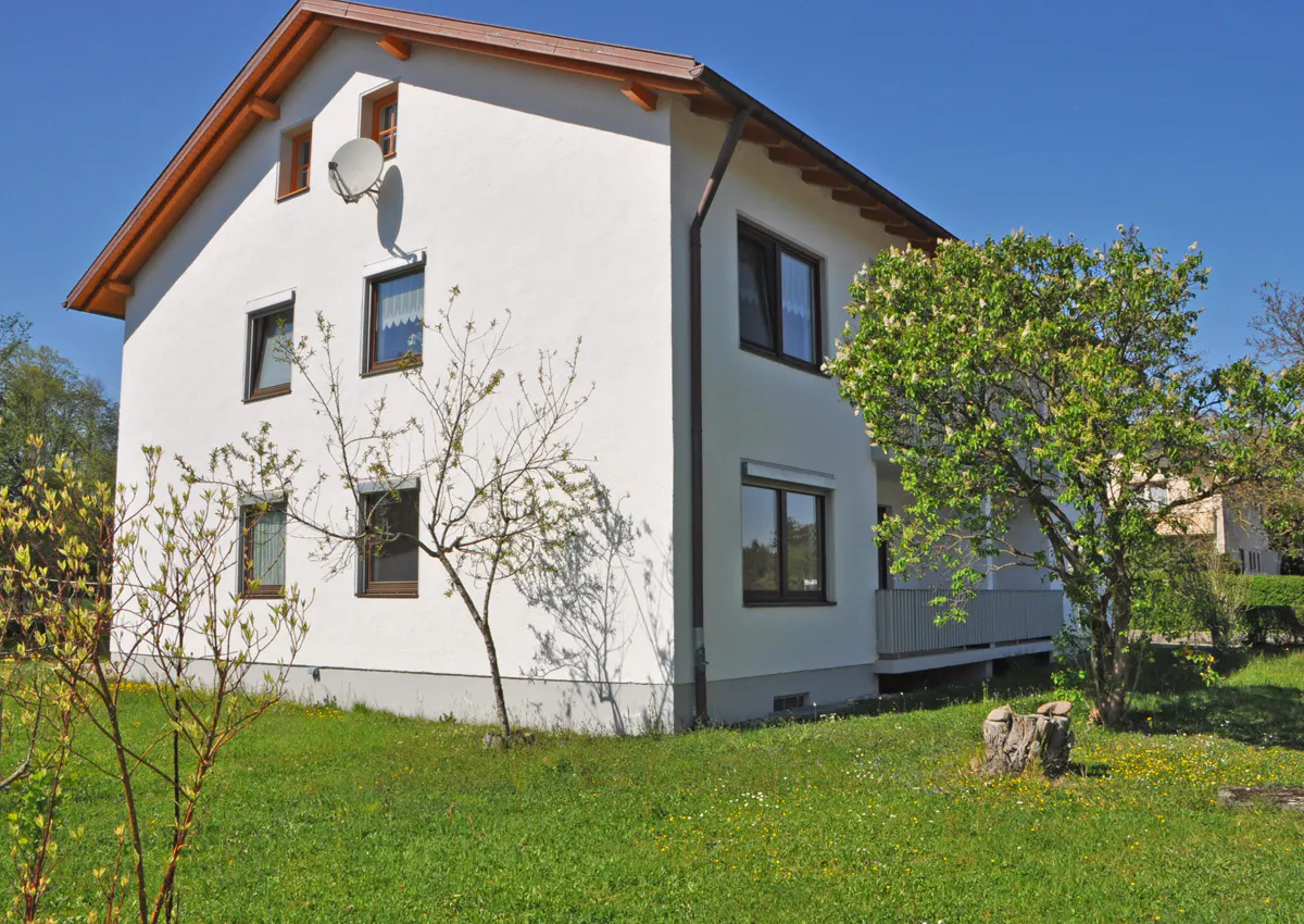 Two-story white house with brown trim and a satellite dish on the side, surrounded by green grass and trees under a blue sky.