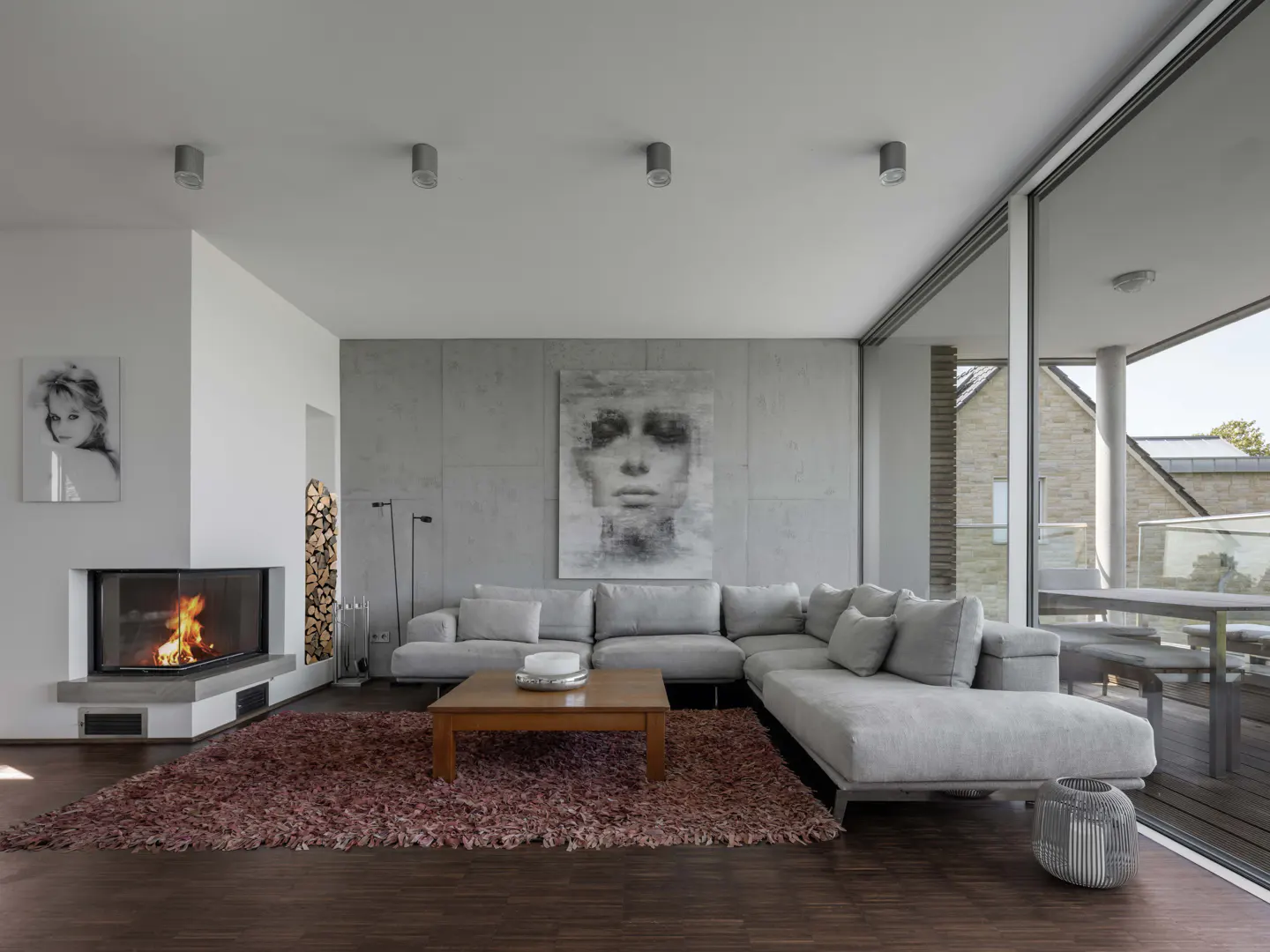 Living room with gray sectional sofa, wood coffee table, and a fireplace with burning logs. Large windows offer outdoor views.