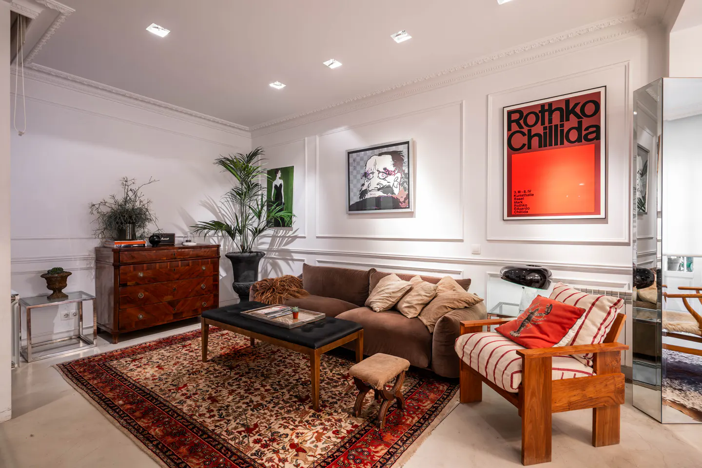 Living room with brown sofa, wooden chair, and patterned rug. Art adorns the white walls. A chest of drawers sits near a potted plant.