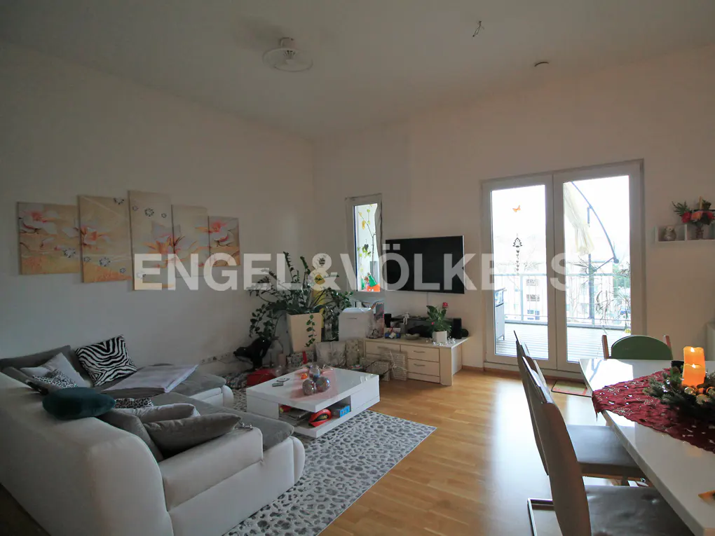 Bright living room with a white sectional sofa, coffee table, TV, and dining area with light wood floors and balcony doors.