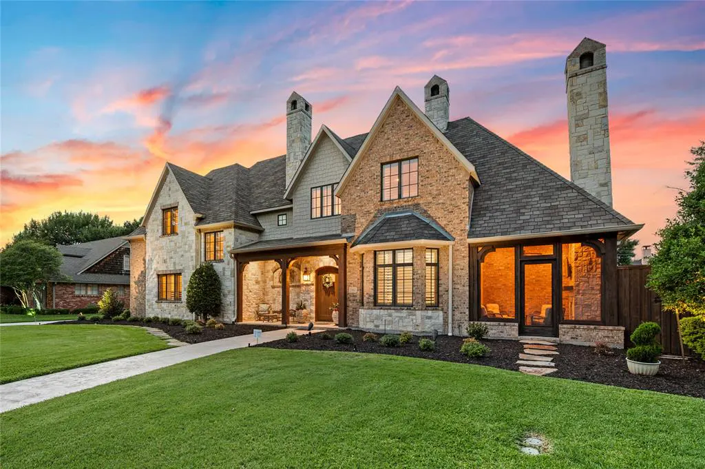 Luxury stone and brick home with a covered porch and tall chimneys at sunset, featuring a manicured lawn and stone path.