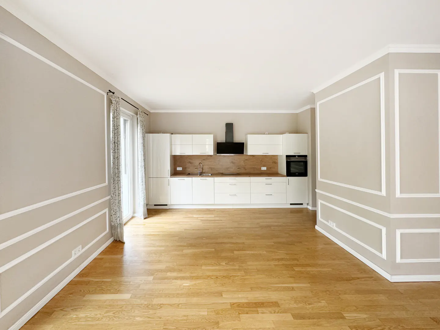 Bright, empty room with wood floors, beige walls with white trim, and a white kitchen with wood backsplash.