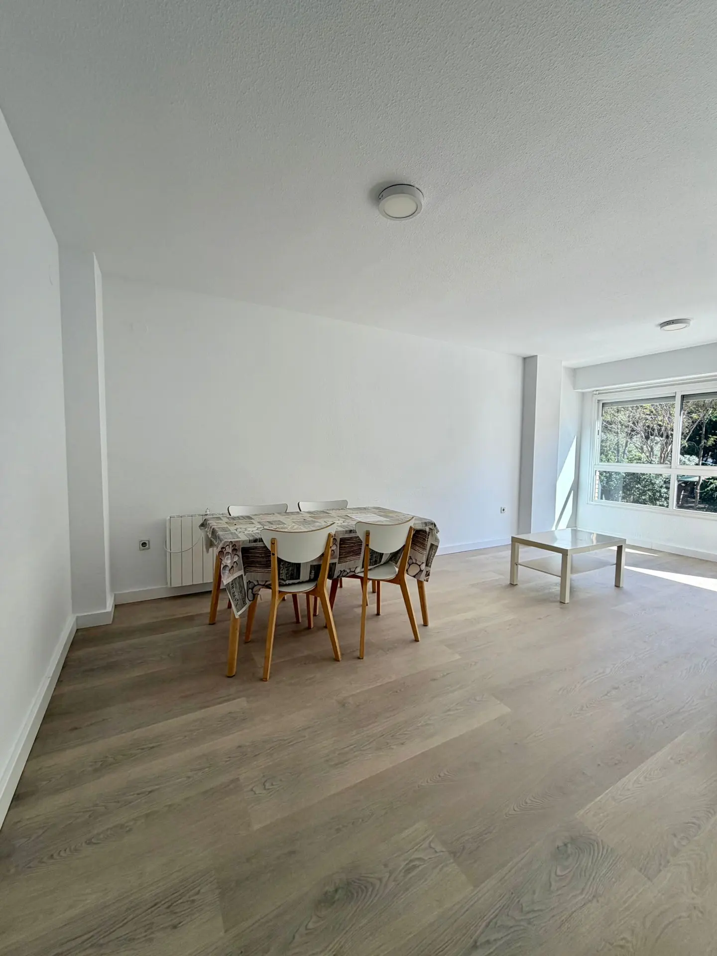 Bright, empty room with wood floors, white walls, and a large window. A dining table with chairs and a coffee table are present.