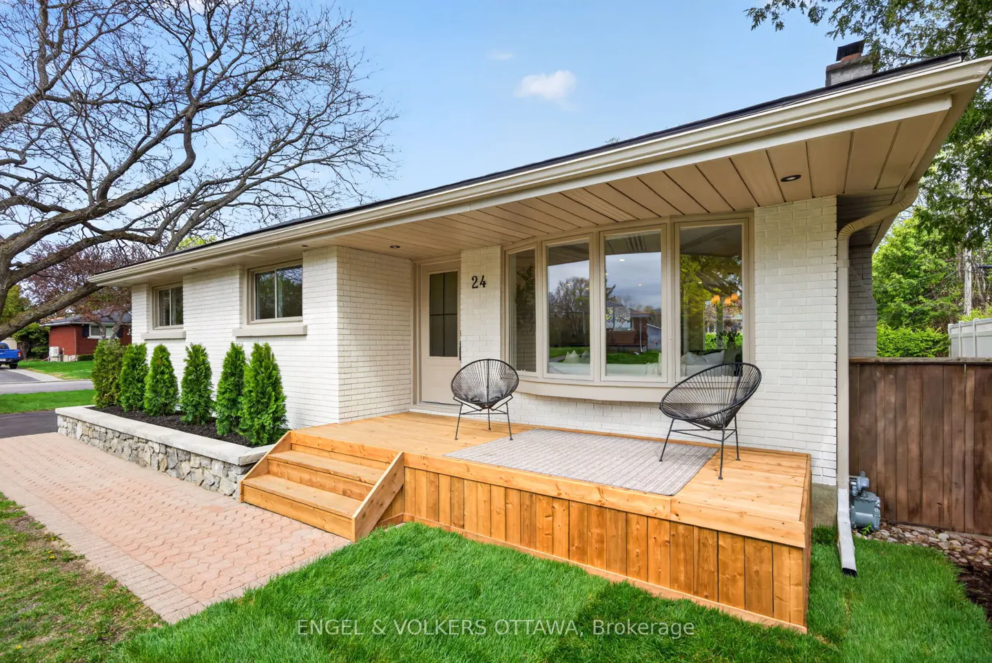 Exterior view of a white brick house with a wooden porch, two black chairs, and green lawn.