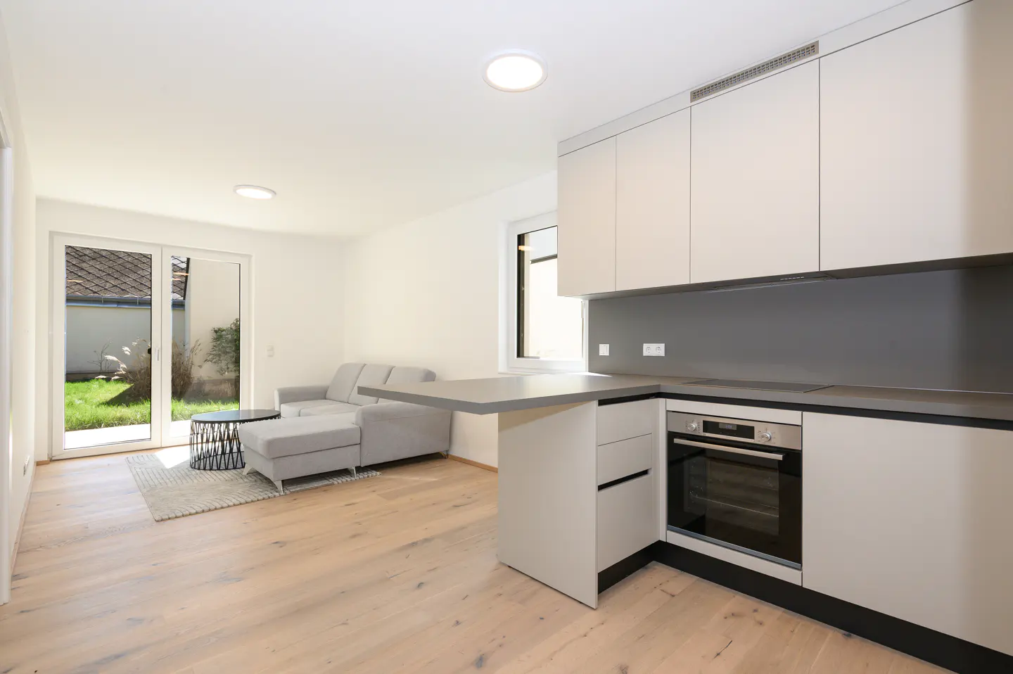Bright, modern apartment with light wood floors. A gray sofa faces sliding glass doors to a green yard. White cabinets and a gray countertop define the kitchen.