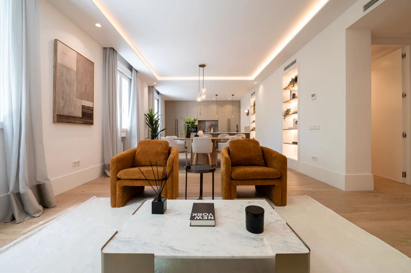 Bright, modern living room with two orange armchairs, a marble coffee table, and an open-concept kitchen and dining area in the background.