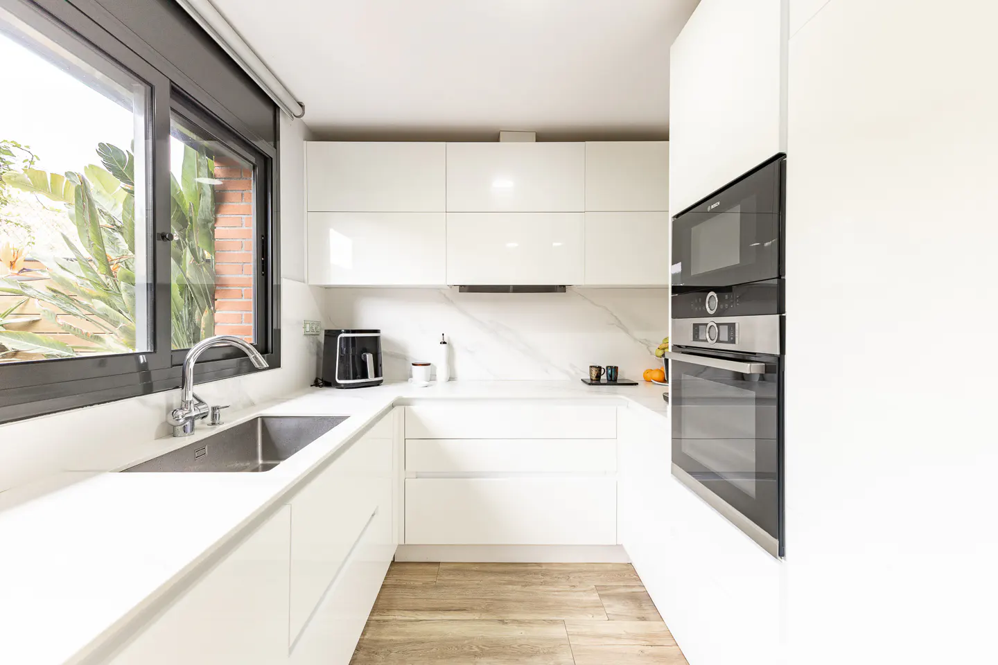 Bright, modern kitchen with white cabinets, marble countertops, and stainless steel appliances. A window overlooks lush greenery.