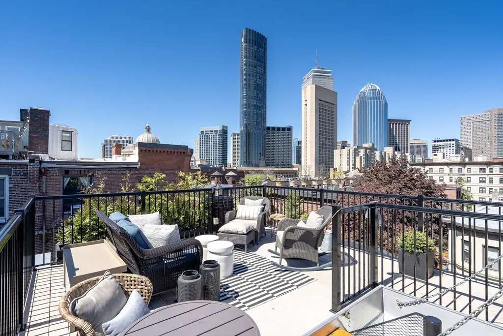 Rooftop patio with wicker furniture, black and white striped rug, and a view of the Boston skyline on a clear day.