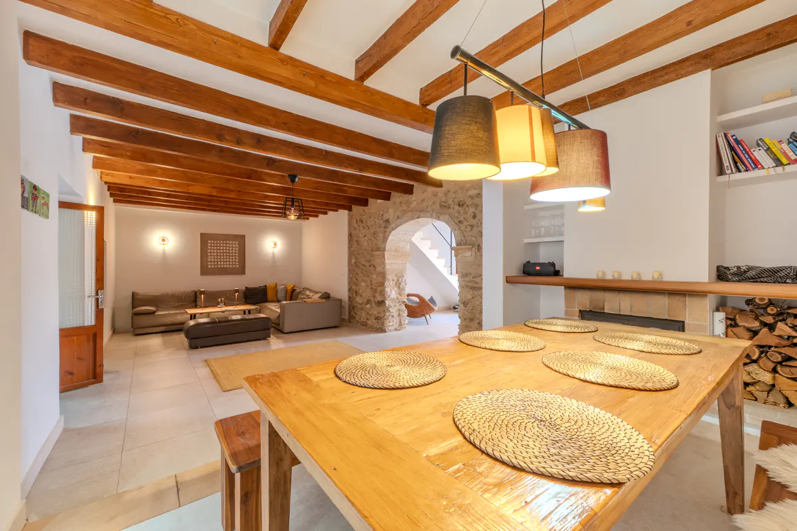 Rustic dining area with wooden table, woven placemats, and exposed ceiling beams, opening into a cozy living room.