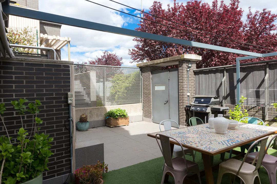 Outdoor patio with a mosaic table, metal chairs, grill, and brick walls. Red-leaved trees are visible in the background.