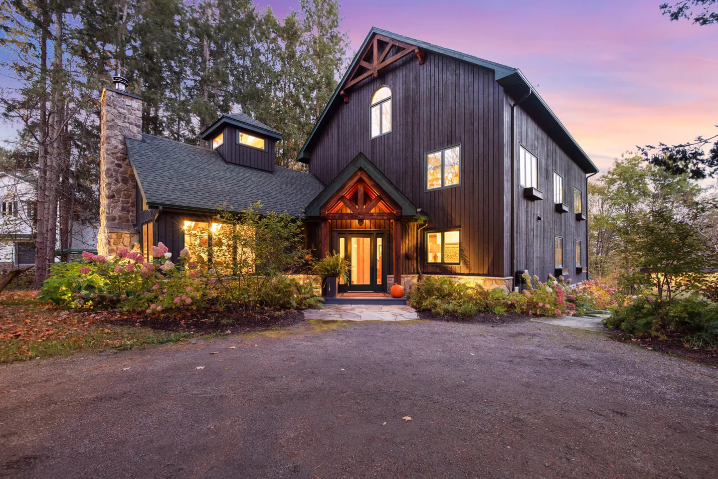 A two-story dark brown house with a stone chimney and green roof, surrounded by trees and pink hydrangeas.