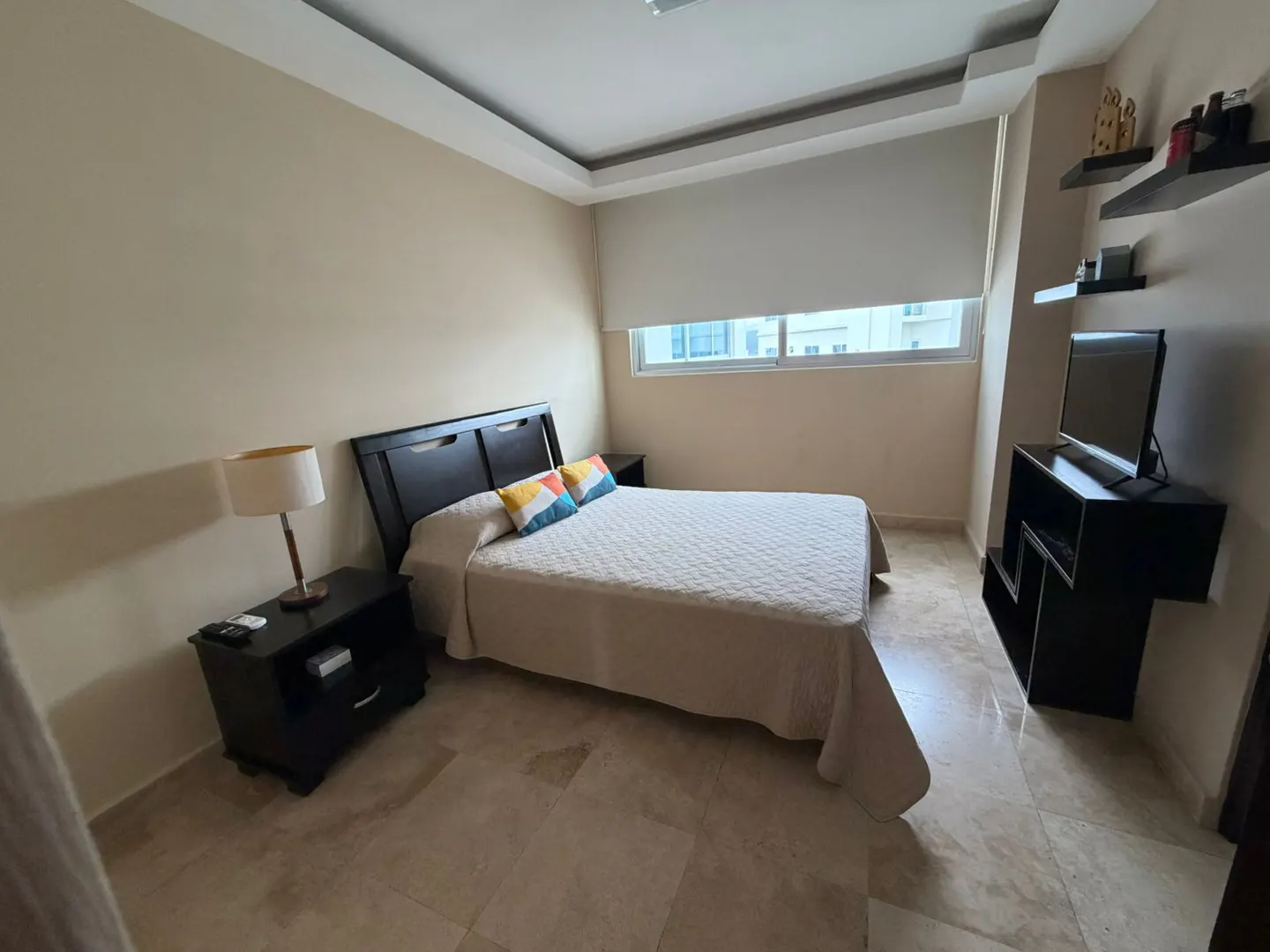 Modern bedroom with a queen bed, dark wood furniture, beige walls, tile floors, and a window with a white roller shade.