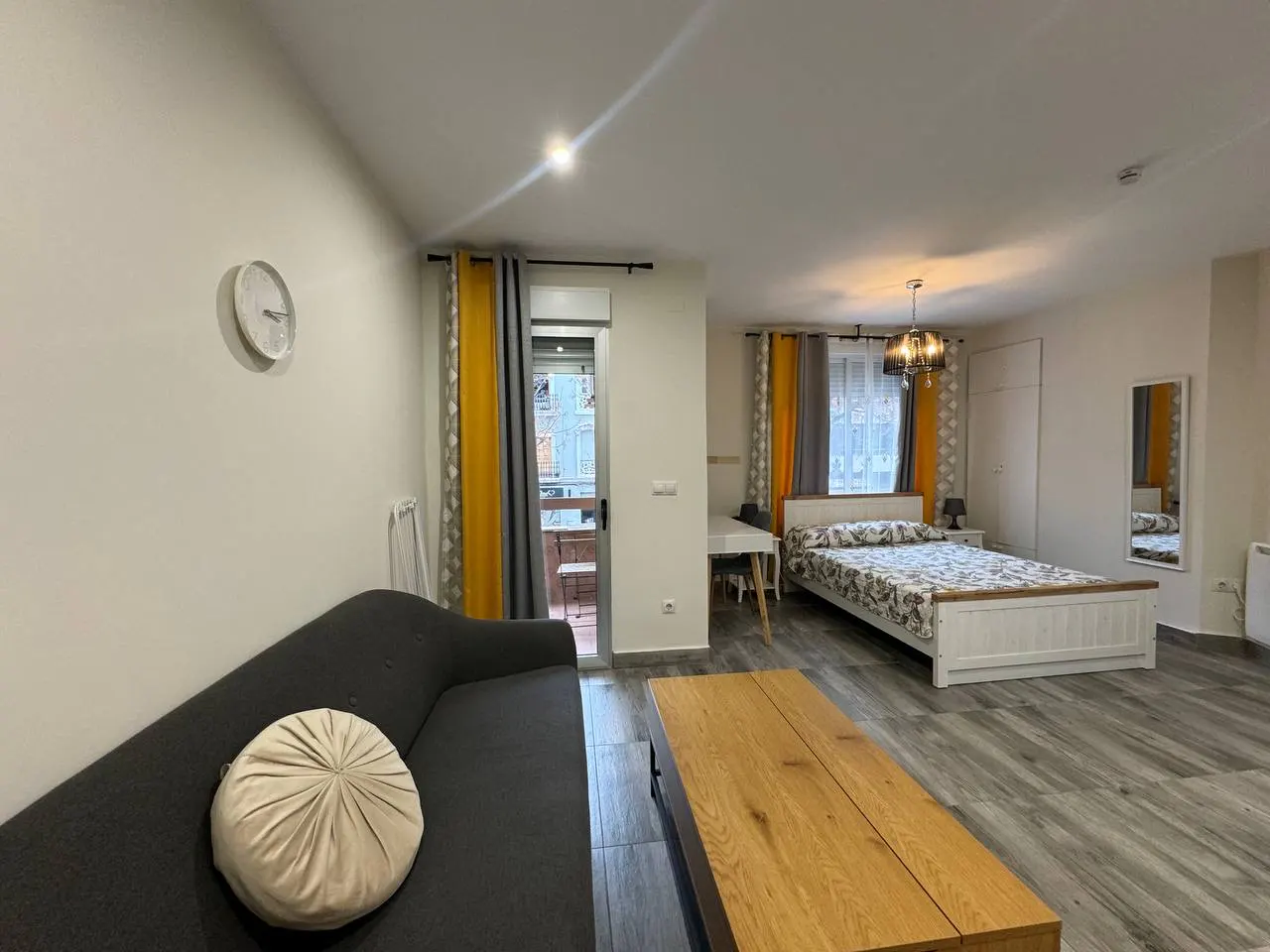Studio apartment with a gray sofa, wooden coffee table, bed, desk, and yellow and gray curtains.
