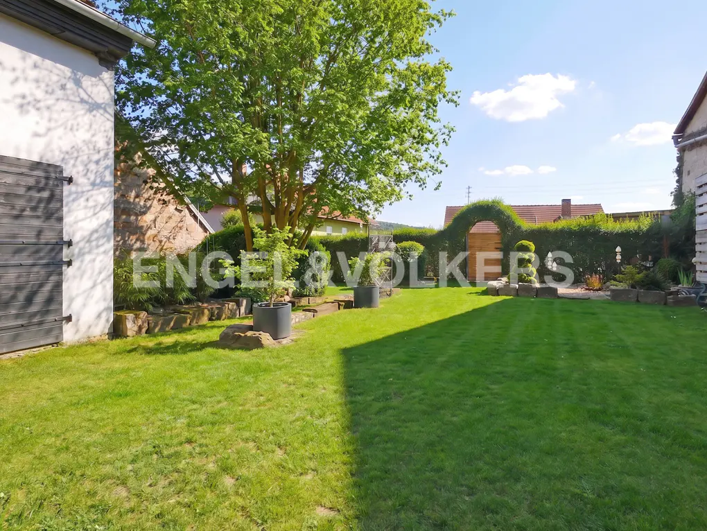 Lush green lawn with trees, shrubs, and stone borders. A hedge archway leads to a wooden gate. Blue sky with clouds.