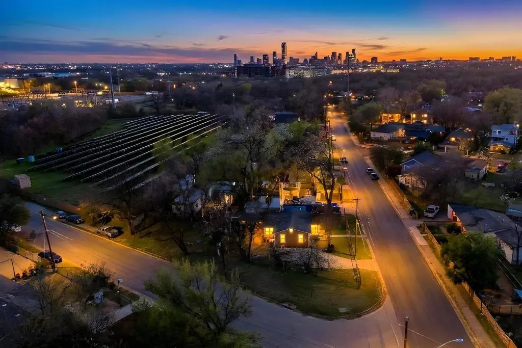 Aerial view of a neighborhood street at dusk, with city skyline in the distance. Solar panels are visible on a nearby hill.