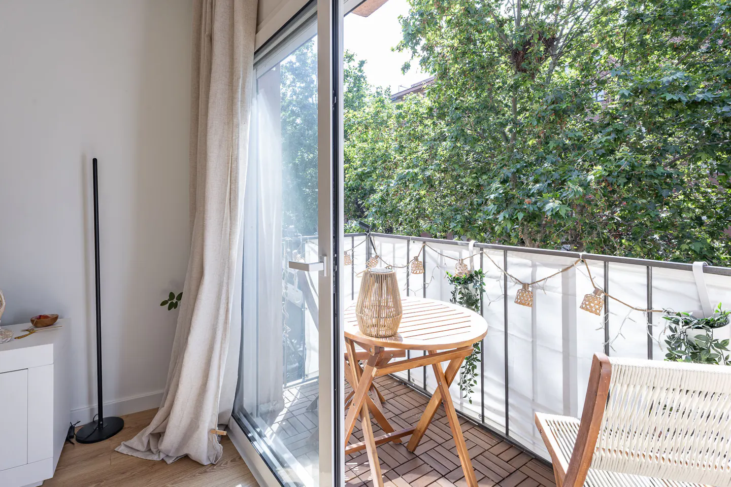 Balcony with a round wooden table, two chairs, and string lights. Green trees are visible outside.