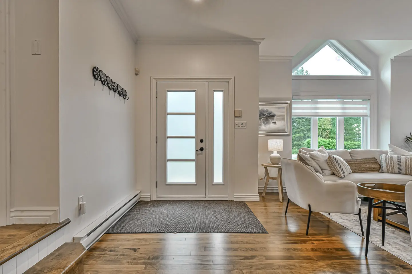 Bright entryway with white walls, wood floors, and a white door with frosted glass. A coat rack hangs on the wall. Living room visible in the background.