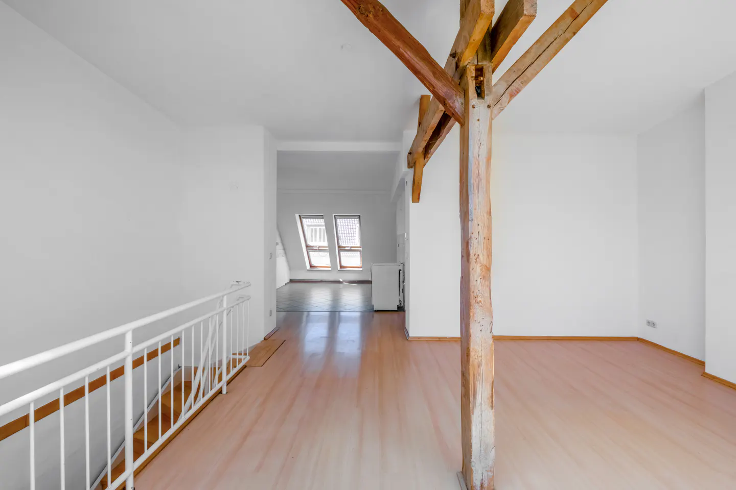 Bright, empty room with light wood floors and white walls. A wooden support beam is in the foreground. Windows are visible in the background.