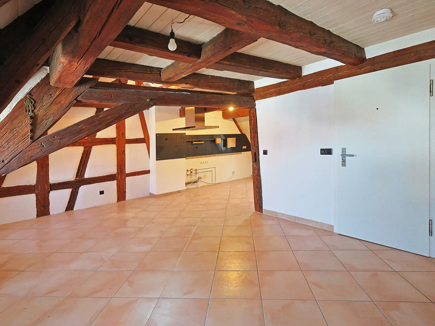 Attic apartment with exposed wood beams, tile floor, and a white door. A kitchen area is visible in the background.