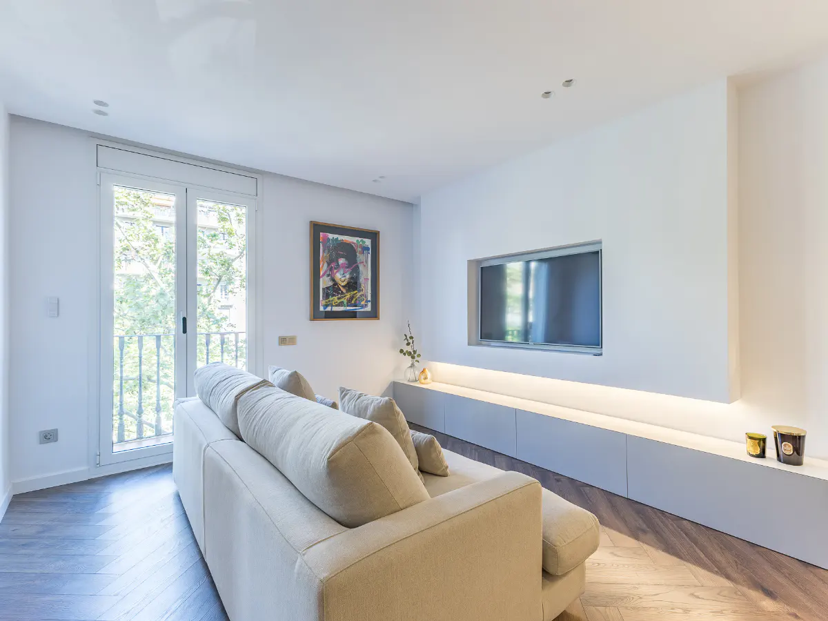 Bright living room with white walls, a beige sofa, and a flat-screen TV mounted above a long, low white cabinet with soft lighting. Balcony doors to the left.
