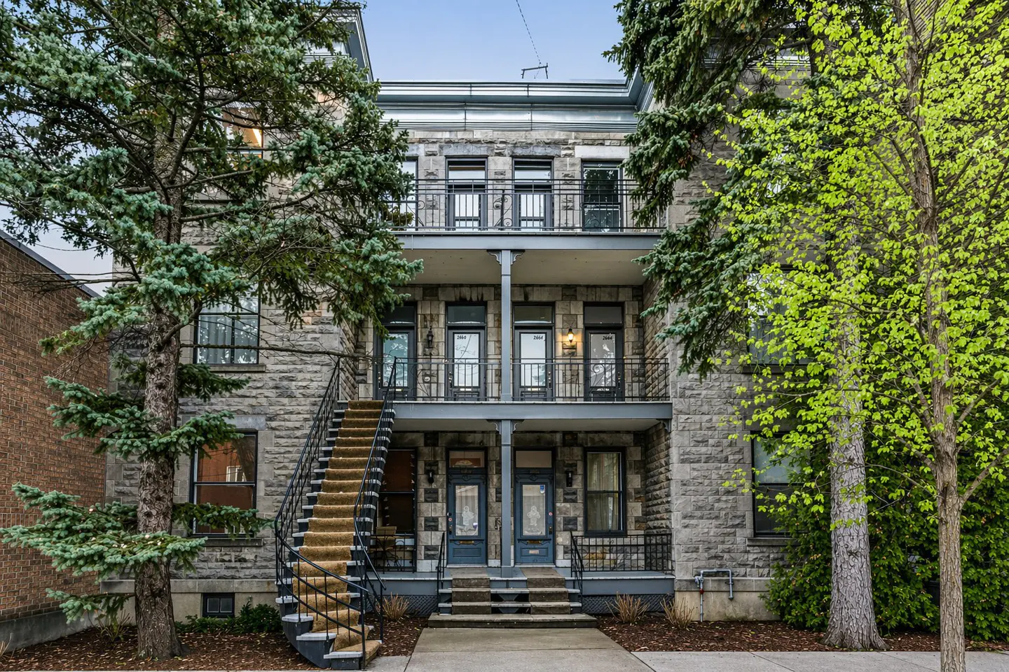 Exterior of a three-story gray stone apartment building with black iron balconies and staircases, framed by green trees.