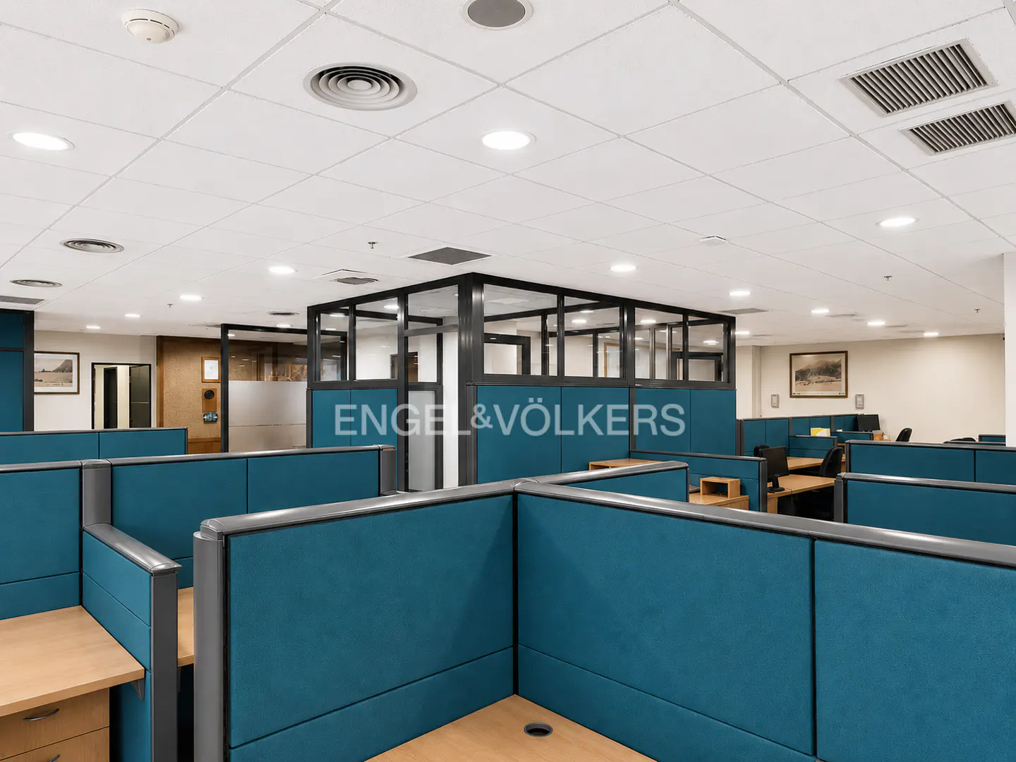 Office space with blue cubicles and a glass-enclosed conference room. The ceiling is white with recessed lighting and vents.
