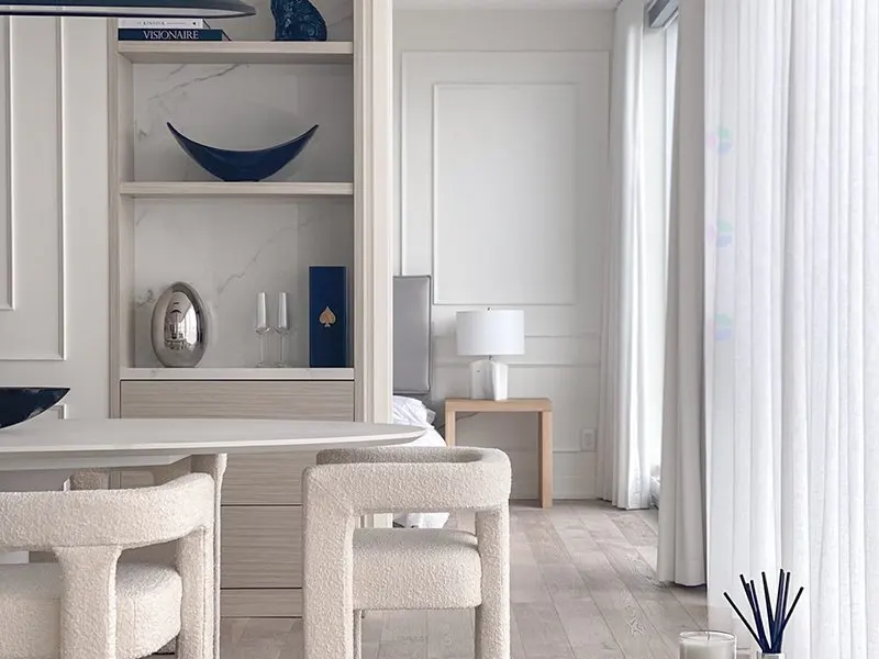 Modern dining area with cream bouclé chairs, a marble-backed shelving unit, and a view into a bright, minimalist bedroom.