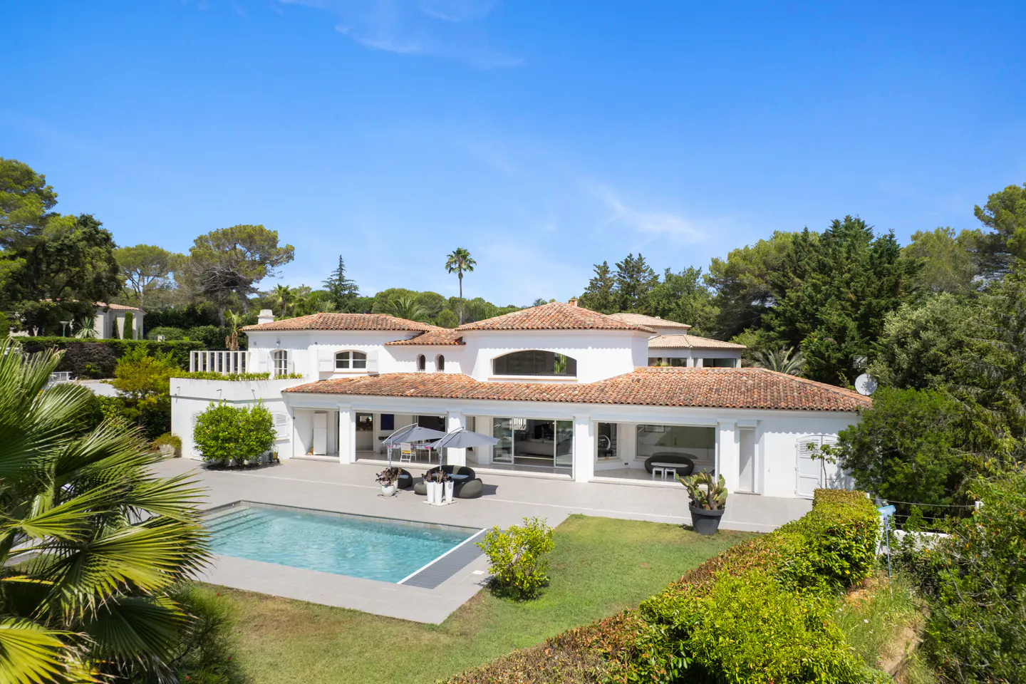 A white villa with a red tile roof, a pool, and green trees under a blue sky.